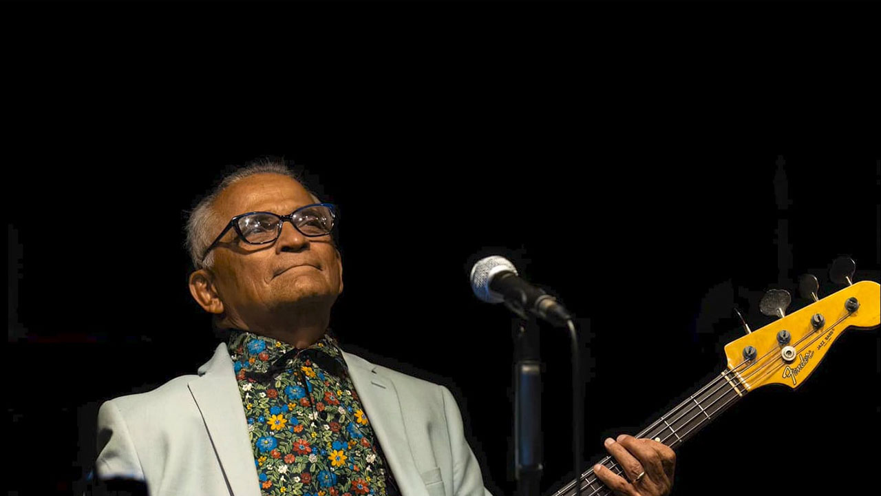Gabriel Espinosa performing on stage with a bass guitar, wearing a light-colored jacket and floral shirt, captured in dramatic lighting