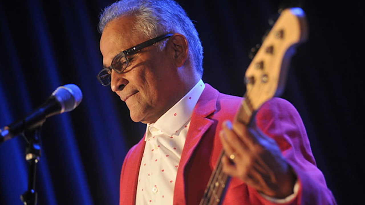 Gabriel Espinosa performing live in a red jacket, playing bass guitar under soft stage lighting
