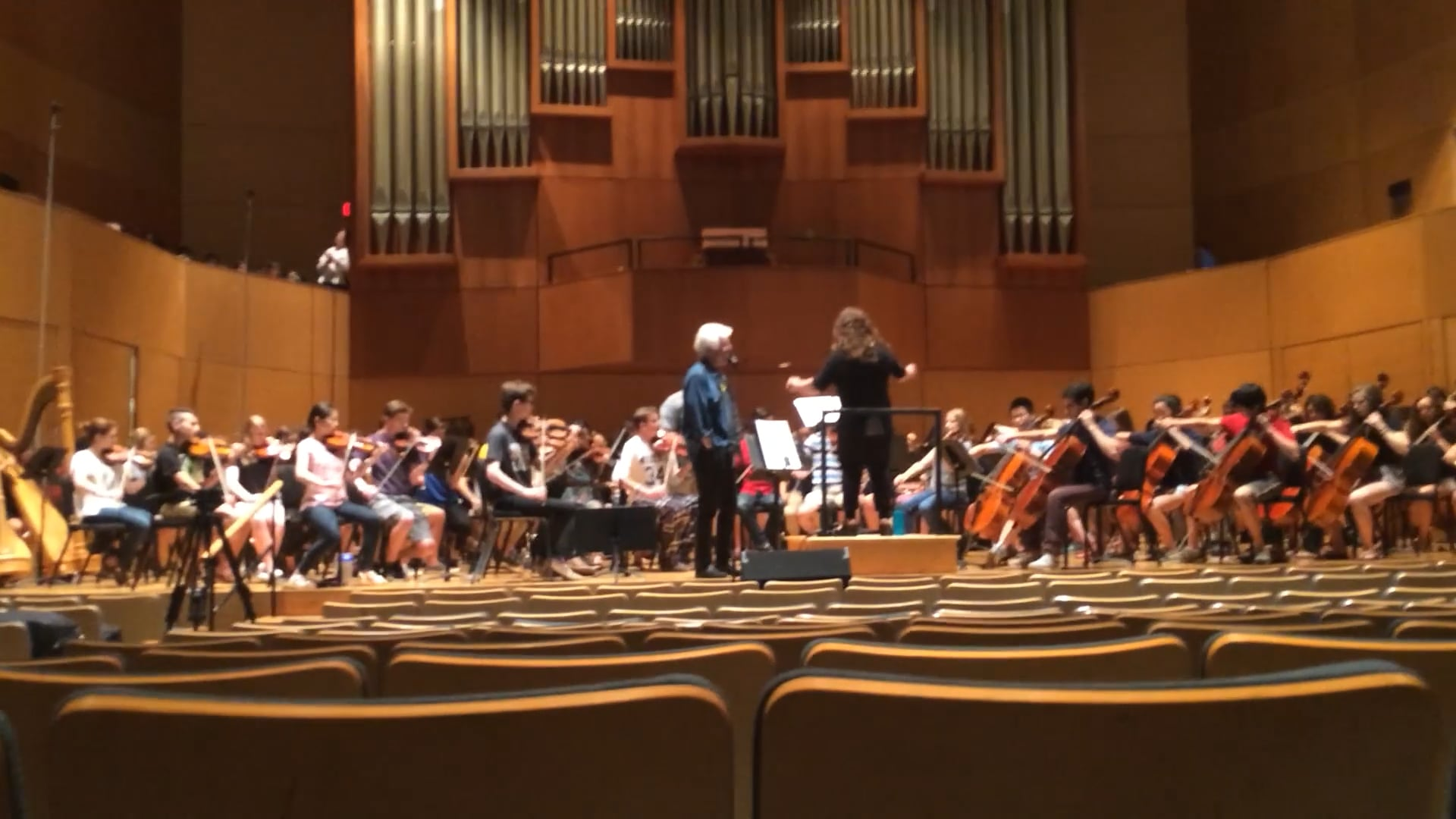 Youth orchestra rehearsing on stage with conductor and guest artist, inside a modern concert hall