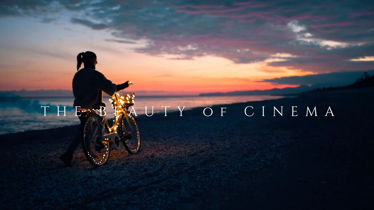 Silhouette of a person walking a bicycle wrapped in lights along a beach at sunset, with text overlay that reads "The Beauty of Cinema"